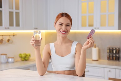 Photo of Happy young woman with glass of water and pills at table in kitchen. Weight loss