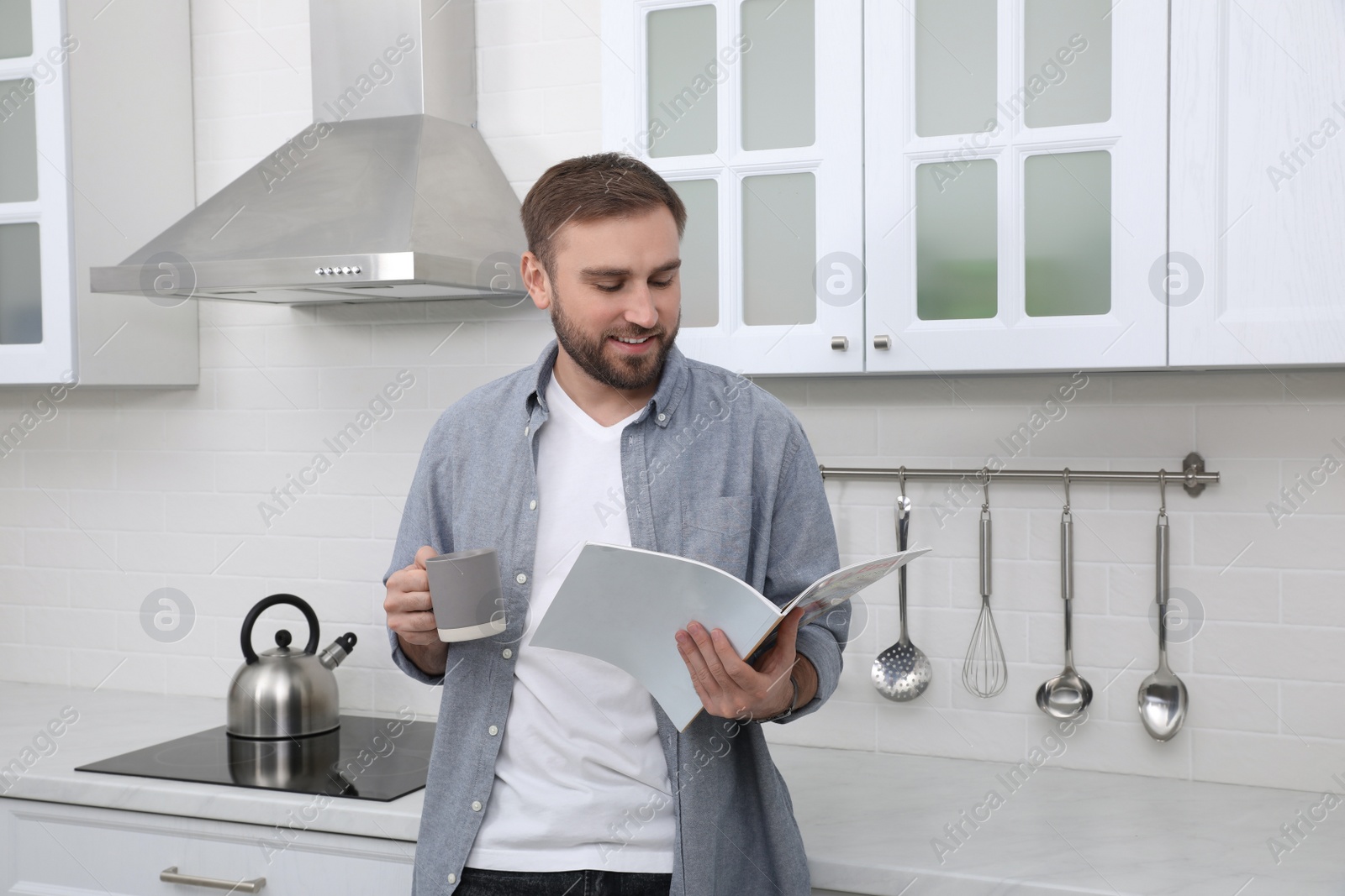 Photo of Handsome man with cup of coffee reading magazine in kitchen