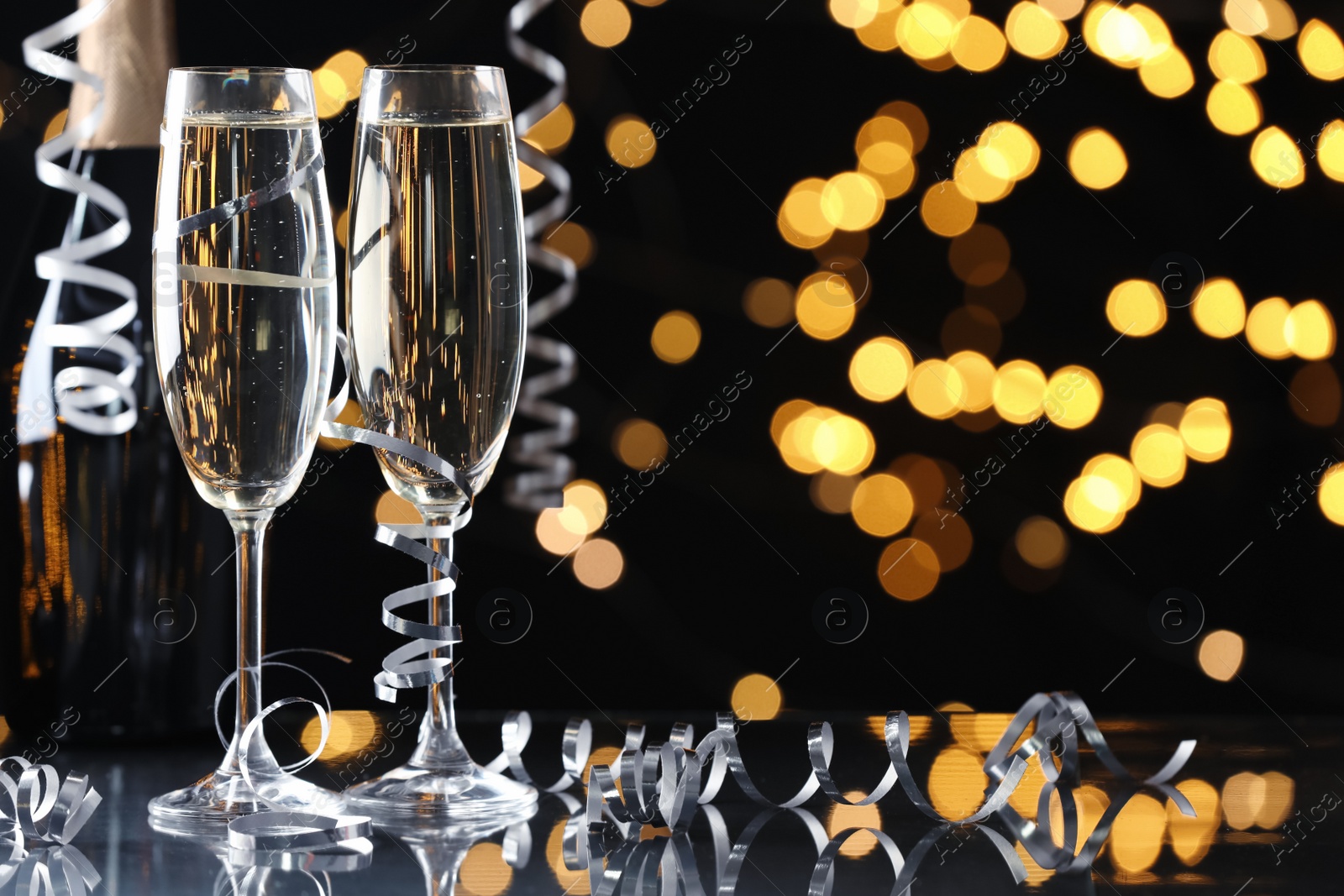 Photo of Glasses and bottle of champagne with serpentine streamers against blurred lights on black background. Space for text
