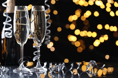 Photo of Glasses and bottle of champagne with serpentine streamers against blurred lights on black background. Space for text