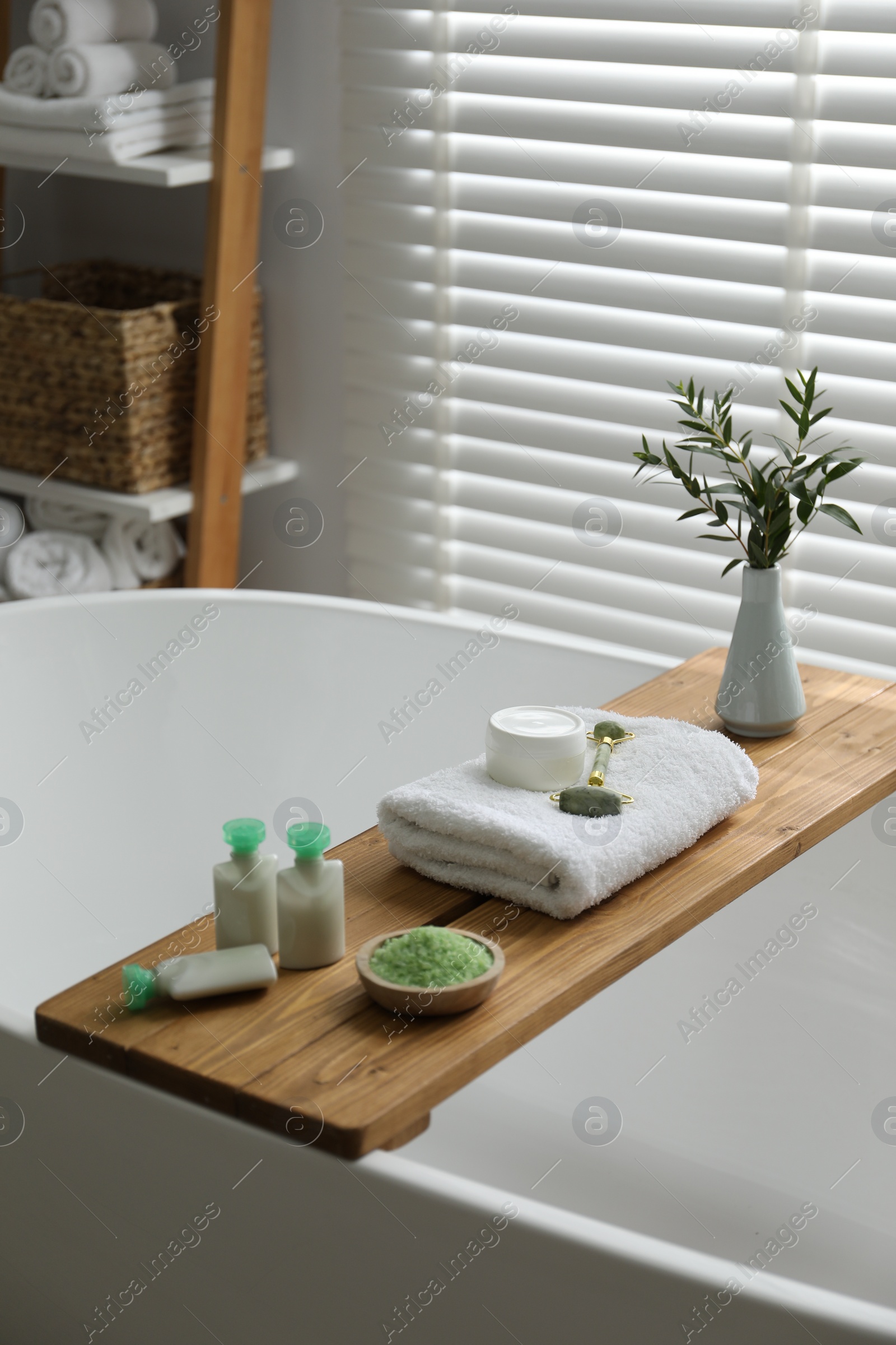 Photo of Wooden tray with spa products and green branches on bath tub in bathroom
