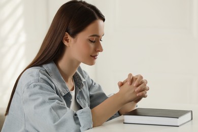 Photo of Religious young woman praying over Bible at table indoors