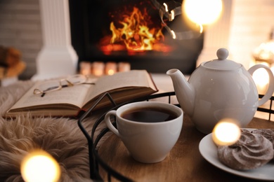 Cup of tea, cookies and book near fireplace indoors. Cozy atmosphere