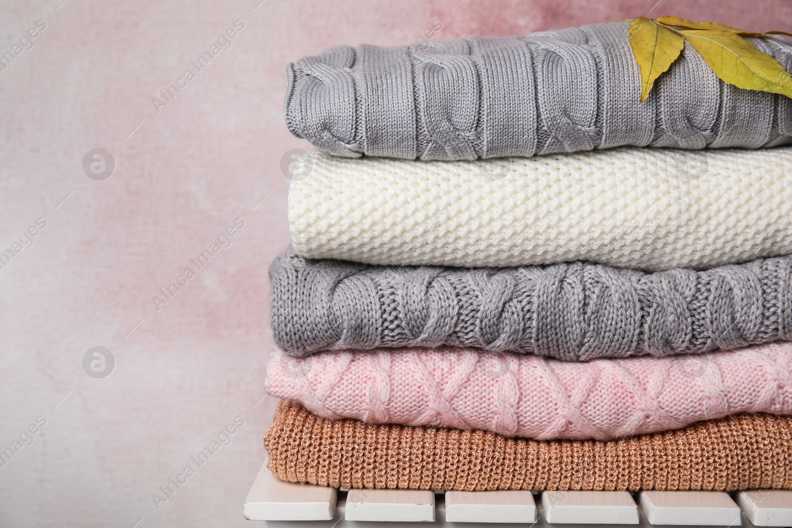 Photo of Stack of warm knitted clothes and autumn leaves on white wooden table against pink background