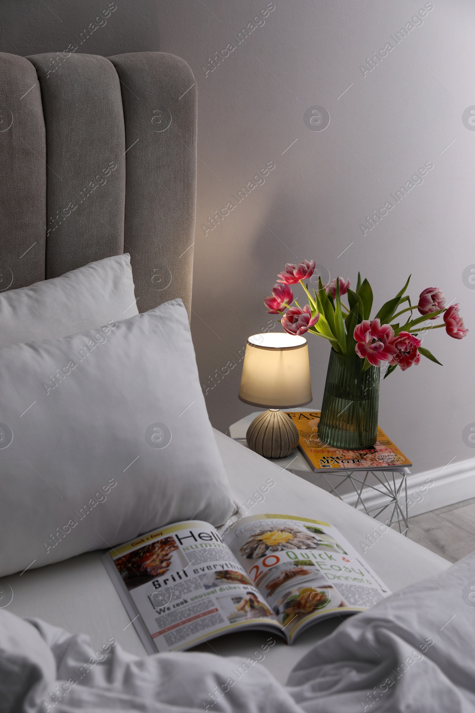 Photo of Stylish lamp, flowers and magazine on bedside table indoors. Bedroom interior elements