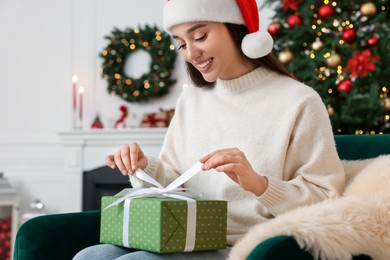 Photo of Happy young woman in Santa hat opening gift box in room decorated for Christmas