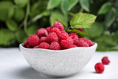 Photo of Bowl of fresh ripe raspberries with green leaves on white table against blurred background, closeup