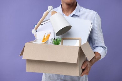 Unemployed young man with box of personal office belongings on purple background, closeup