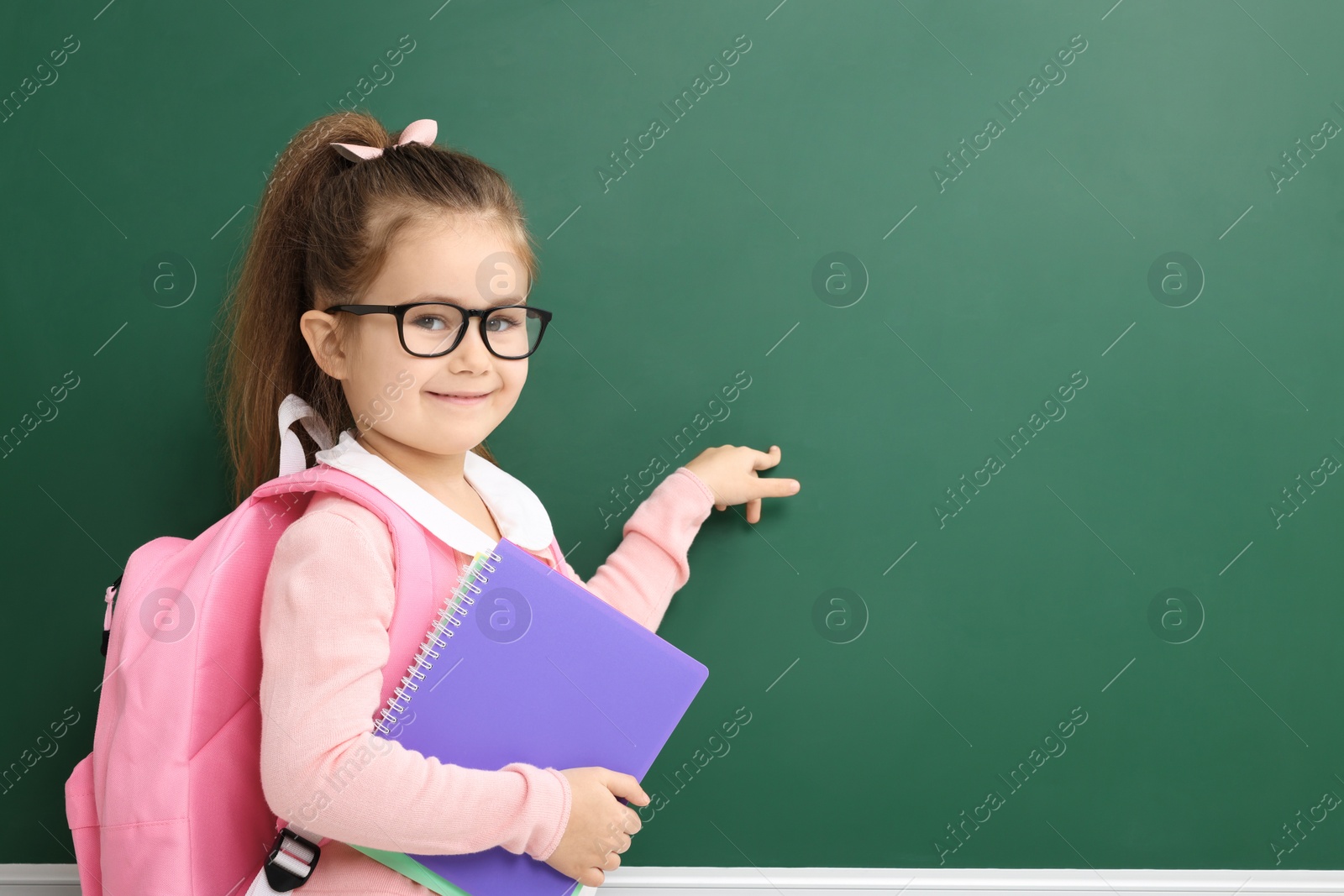 Photo of Happy little school child with notebooks pointing at chalkboard. Space for text