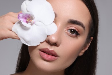 Photo of Beautiful young woman covering eye with orchid flower on grey background, closeup