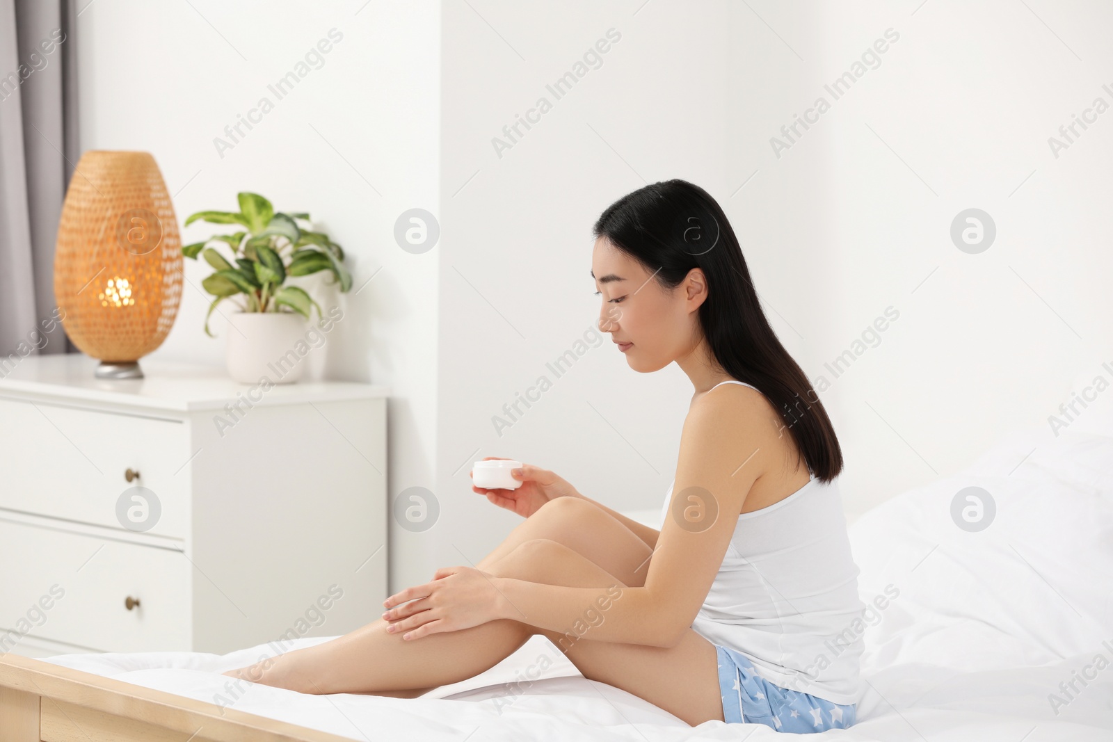 Photo of Beautiful young Asian woman applying body cream on leg in bedroom