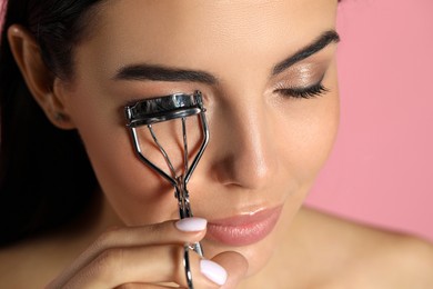 Young woman using eyelash curler on light pink background, closeup