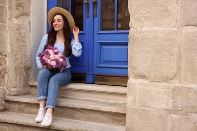 Beautiful woman with bouquet of spring flowers on stairs near building, space for text