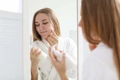 Photo of Young woman with acne problem holding bottle with cosmetic product in bathroom