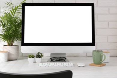 Office workplace with computer, glasses, cup and houseplants on light table near white brick wall