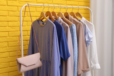 Photo of Rack with different stylish clothes and bag near yellow brick wall in room