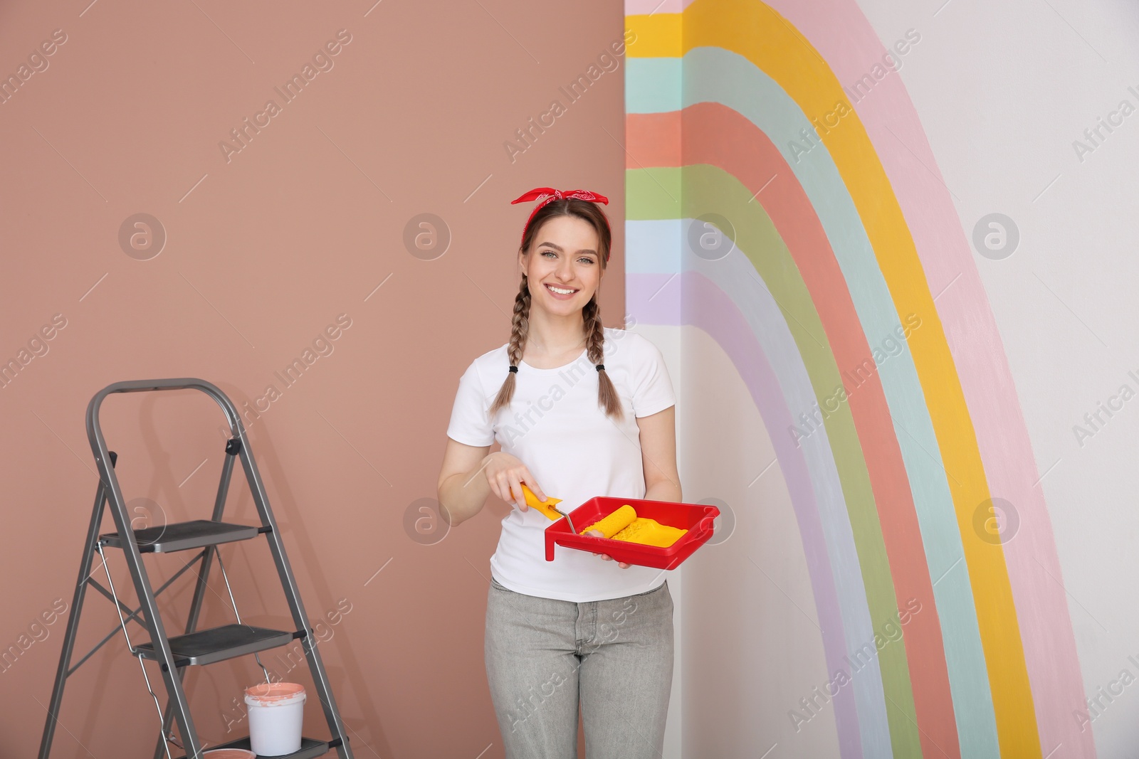 Photo of Young woman holding tray and roller near wall with painted rainbow indoors