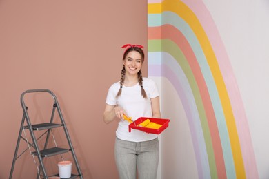 Photo of Young woman holding tray and roller near wall with painted rainbow indoors