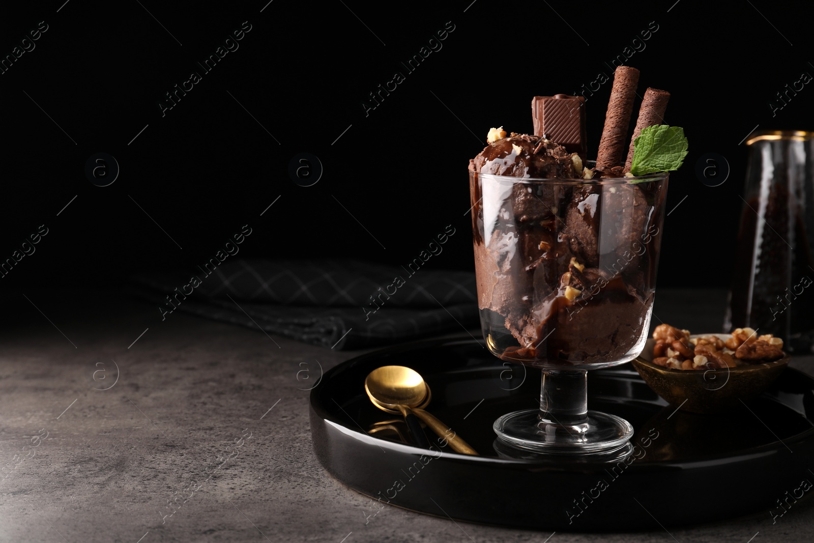 Photo of Tasty chocolate ice cream with sauce, nuts and wafer rolls in glass dessert bowl on dark grey table. Space for text