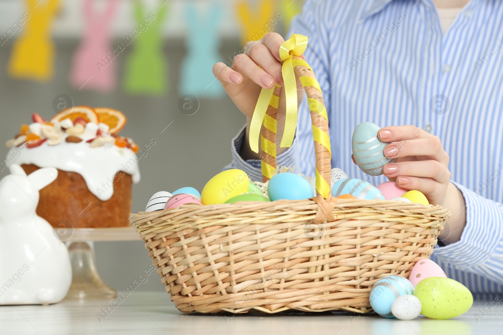 Photo of Woman putting painted egg into Easter basket at table, closeup