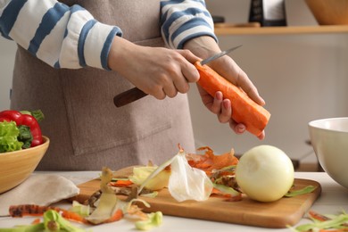 Photo of Woman peeling fresh carrot with knife at table in kitchen, closeup