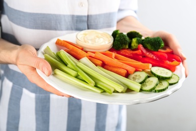 Woman holding plate with celery sticks, other vegetables and dip sauce on light background, closeup