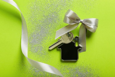 Key with trinket in shape of house, bow, ribbon and glitter on light green background, flat lay. Housewarming party