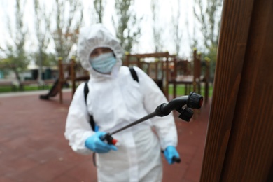 Photo of Woman wearing chemical protective suit at playground, focus on disinfectant sprayer. Preventive measure during coronavirus pandemic