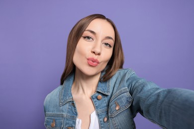 Beautiful young woman blowing kiss while taking selfie on purple background