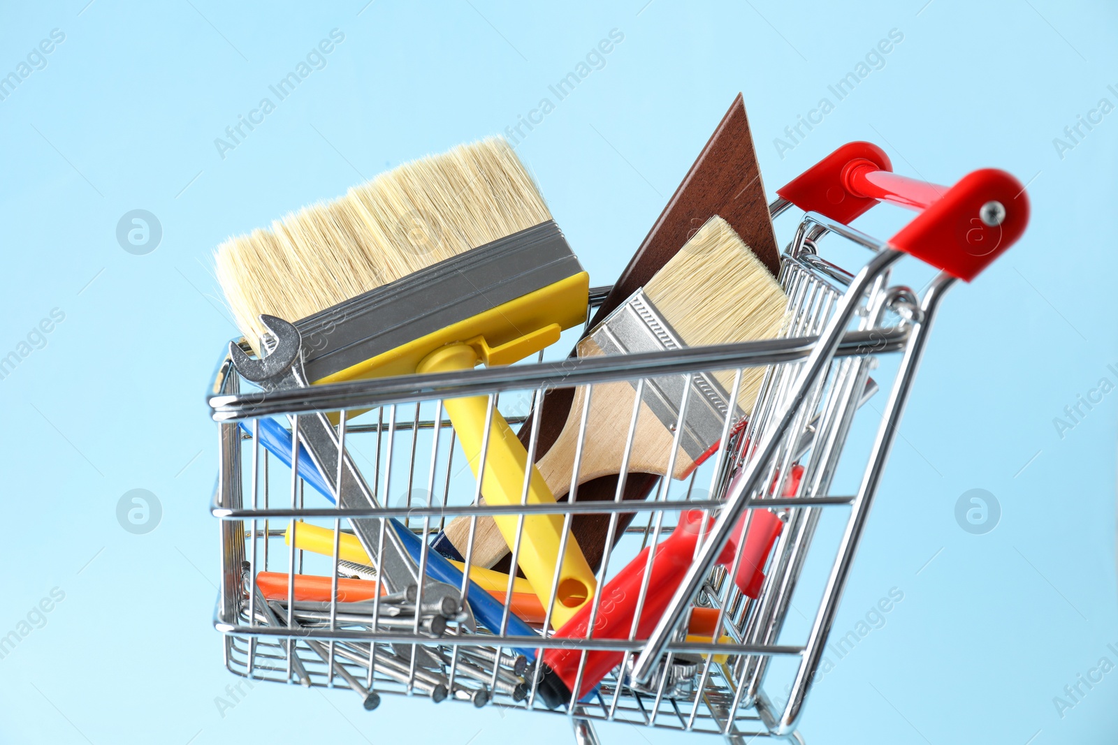 Photo of Different construction tools in shopping cart on light blue background, closeup
