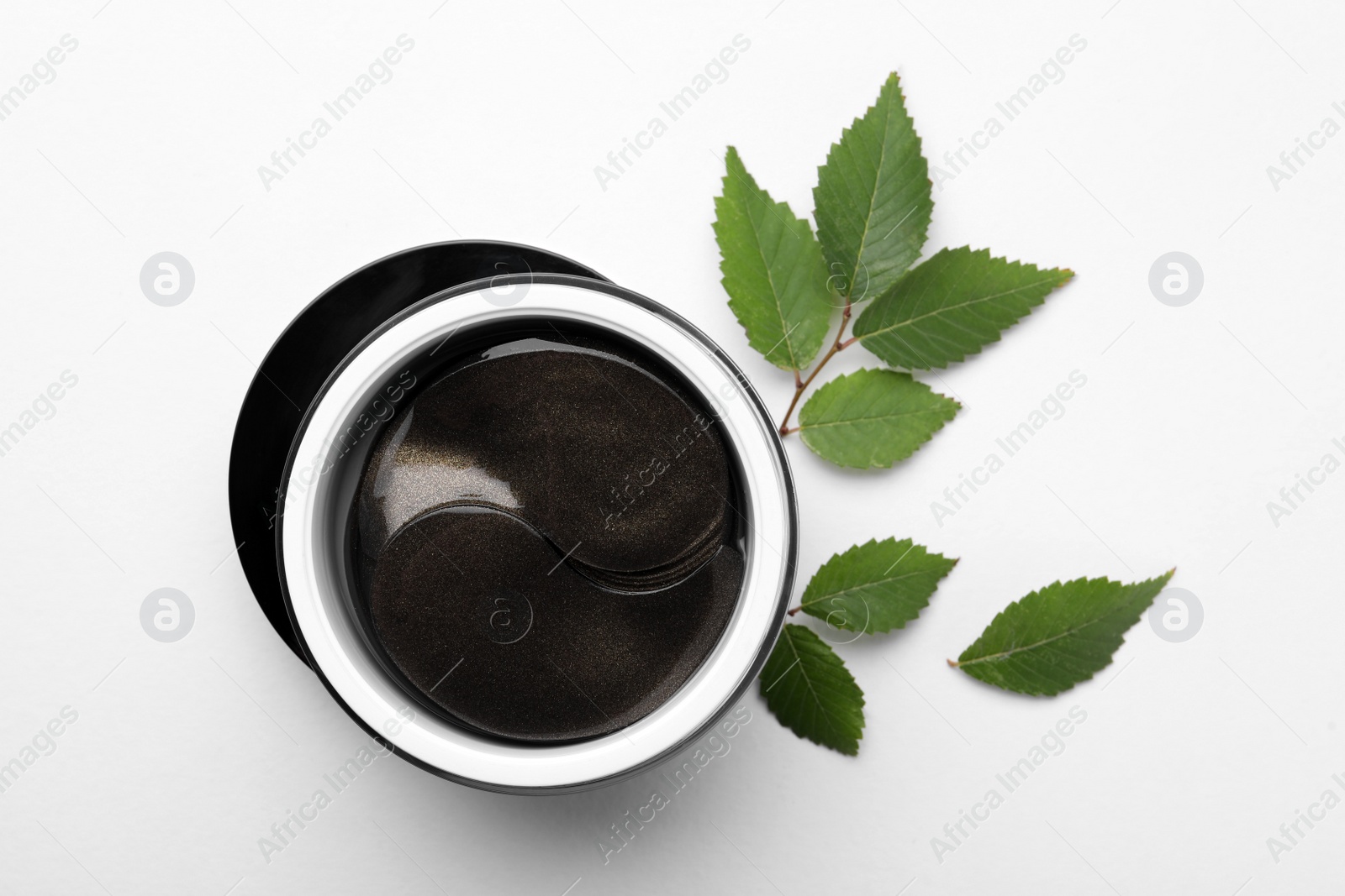 Photo of Under eye patches in jar near green leaves on white background, flat lay. Cosmetic product