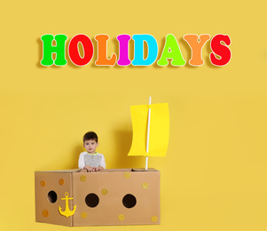 School holidays. Cute little child playing with cardboard ship near yellow wall
