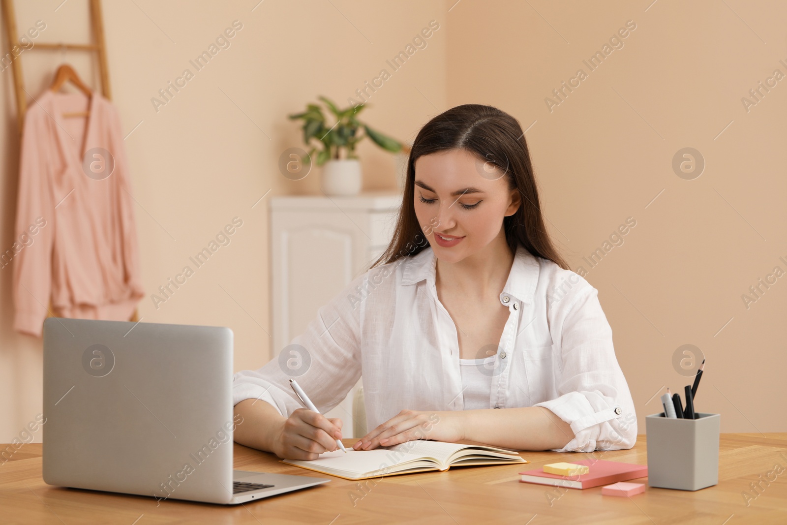 Photo of Happy young woman writing in notebook while working on laptop at wooden table indoors