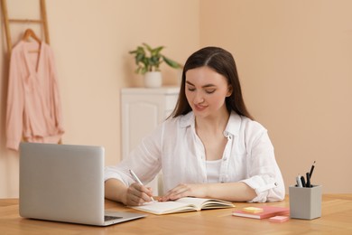 Happy young woman writing in notebook while working on laptop at wooden table indoors