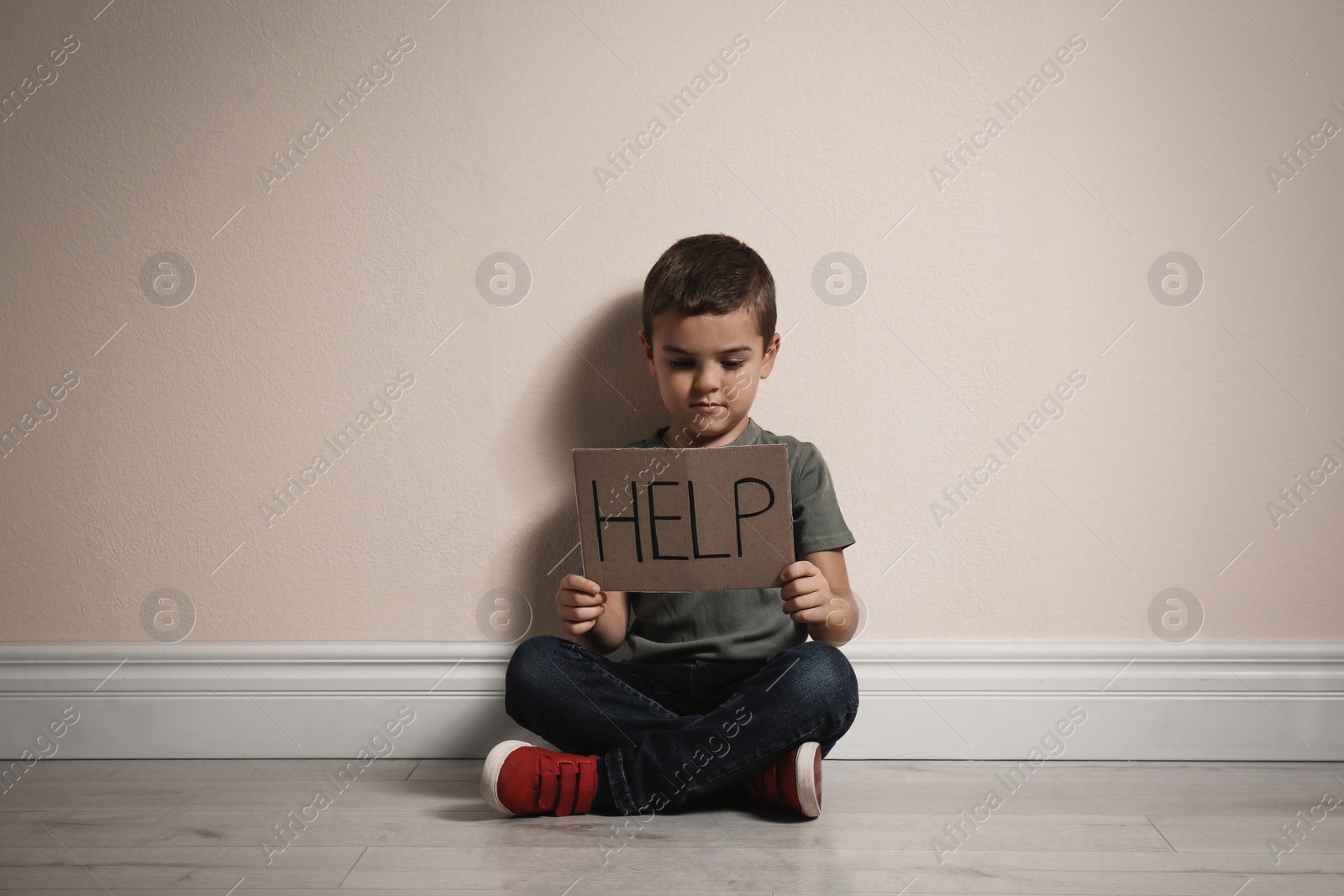 Photo of Little boy with sign HELP sitting on floor near yellow wall. Child in danger