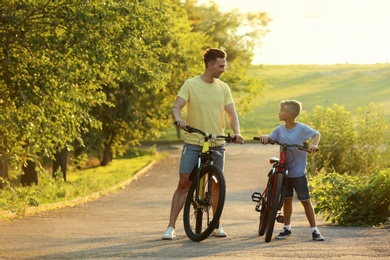 Photo of Dad and son riding bicycles in park on sunny day. Space for text