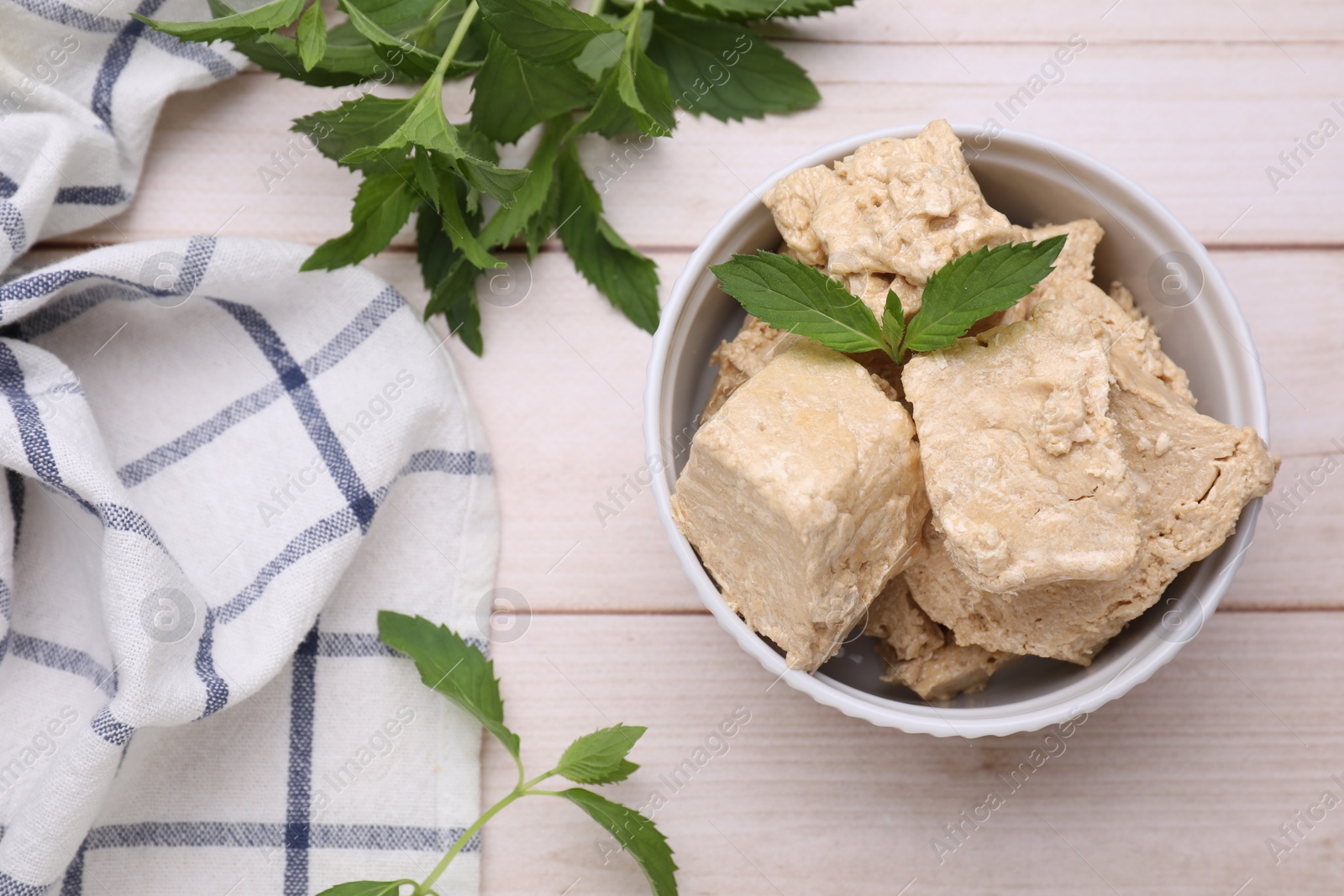 Photo of Bowl with pieces of tasty halva and mint leaves on light wooden table, flat lay