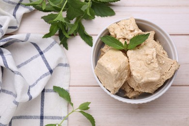 Photo of Bowl with pieces of tasty halva and mint leaves on light wooden table, flat lay