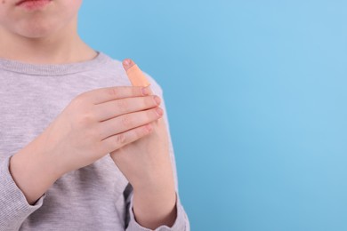 Little boy putting sticking plaster onto finger against light blue background, closeup. Space for text