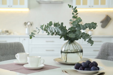 Photo of Beautiful eucalyptus branches,  cups of drink and fresh plums on white table in kitchen. Interior element