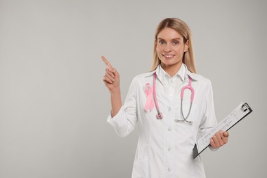 Photo of Doctor with pink ribbon, clipboard and stethoscope on light grey background, space for text. Breast cancer awareness