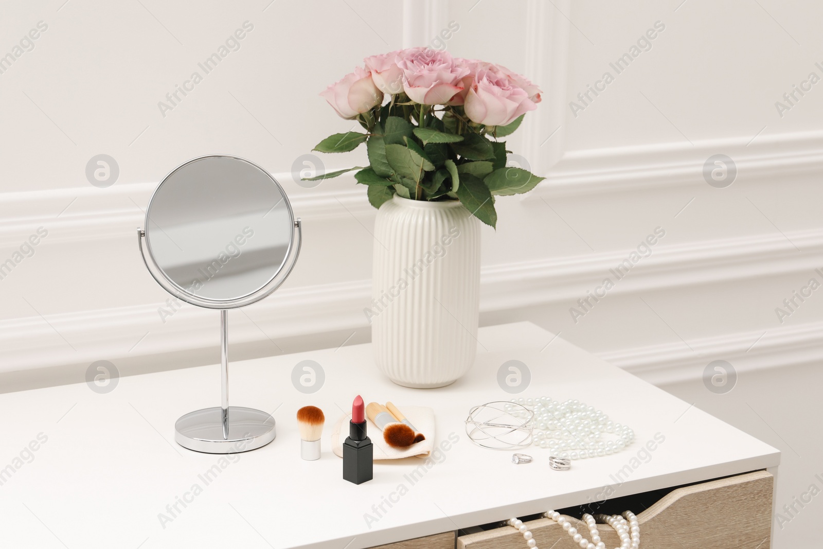 Photo of Mirror, cosmetic products, jewelry and vase with pink roses on white dressing table in makeup room