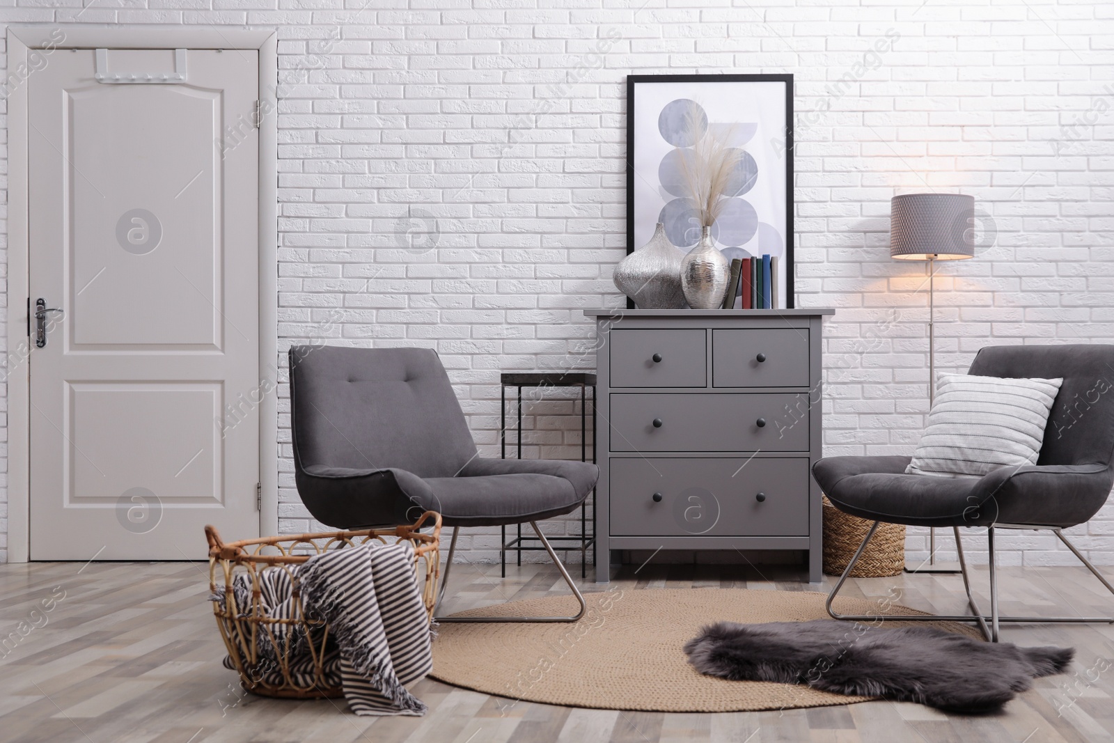 Photo of Stylish room interior with grey chest of drawers and chairs near white brick wall