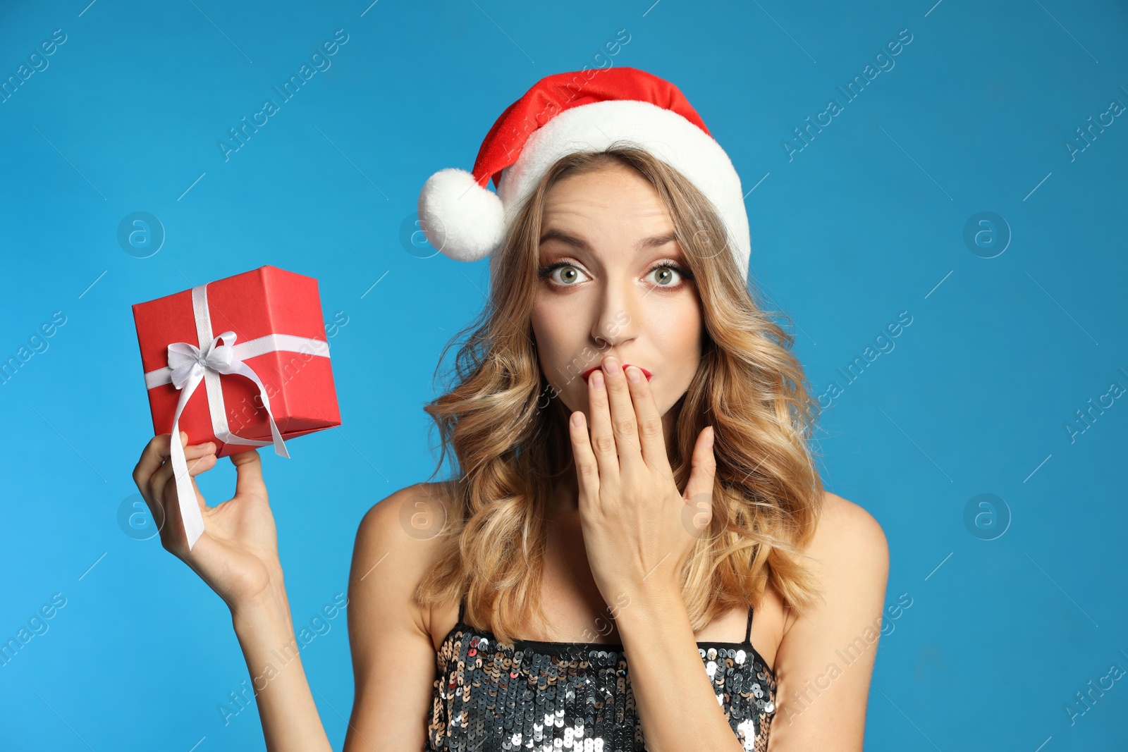 Photo of Surprised young woman wearing Santa hat with Christmas gift on light blue background