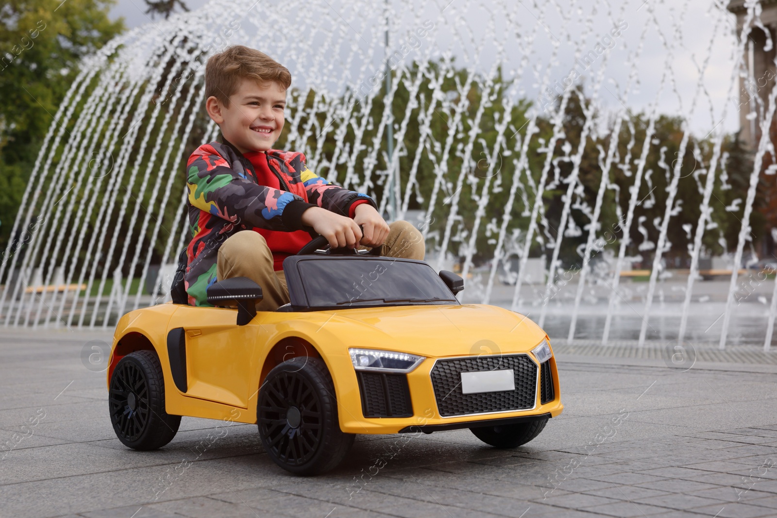 Photo of Cute little boy driving children's car near fountain on city street. Space for text