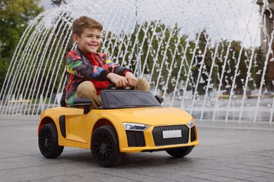 Photo of Cute little boy driving children's car near fountain on city street. Space for text