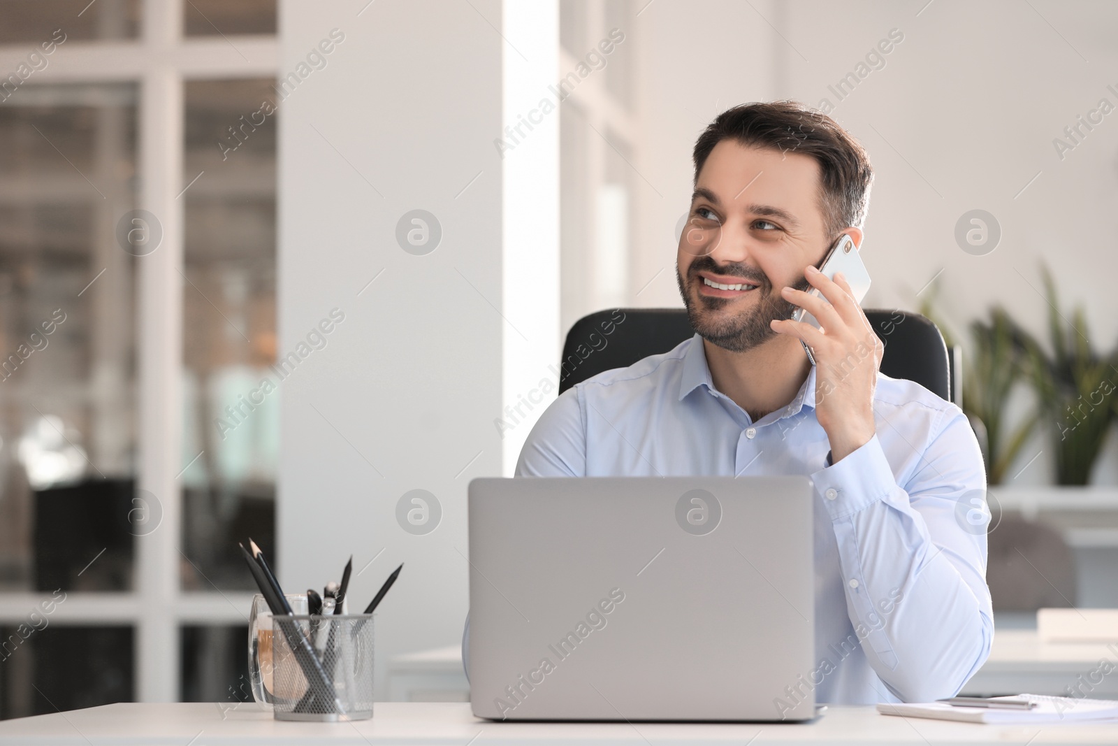 Photo of Happy man using modern laptop while talking on smartphone at white desk in office. Space for text