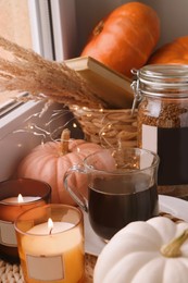 Cup of hot drink, jar with coffee beans, candles and pumpkins on window sill indoors