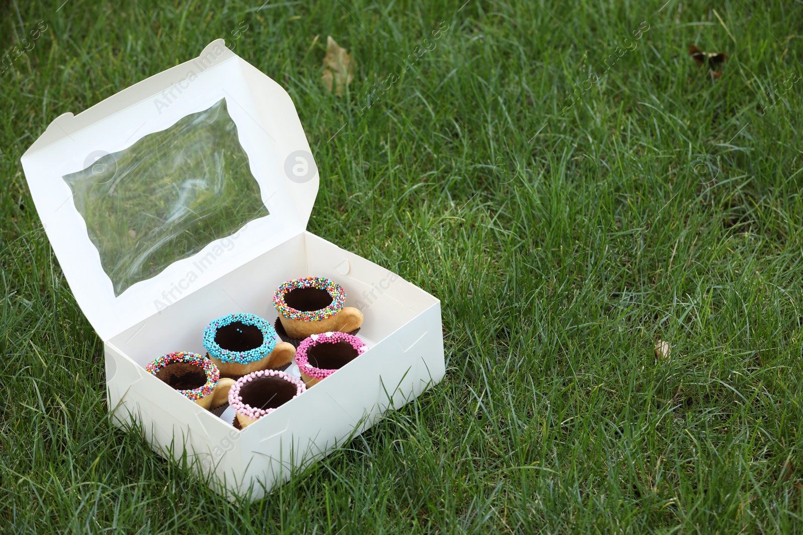 Photo of Box of delicious edible biscuit coffee cups decorated with sprinkles on green grass outdoors, space for text