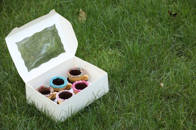 Box of delicious edible biscuit coffee cups decorated with sprinkles on green grass outdoors, space for text
